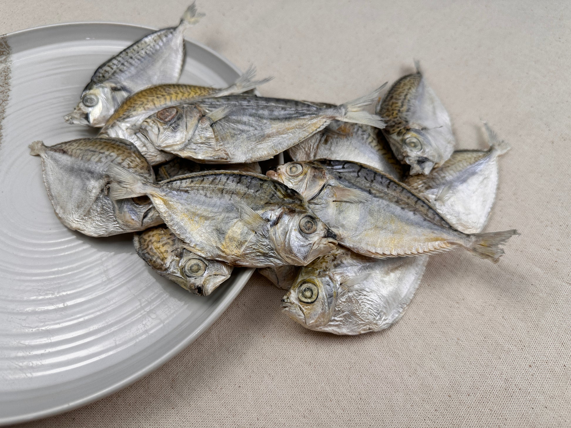 Pile of whole dried ponyfish stacked on a white ceramic plate, displaying their natural texture, eyes, and patterned skin.